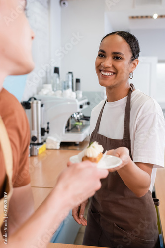 African american seller in apron giving blurred dessert to colleague in confectionery.