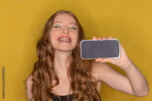 Beautiful cheerful pleased woman with red hair with big lips and braces rejoices and feels happy holding phone on isolated background