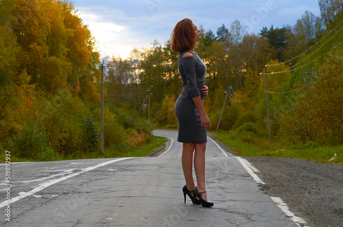a young girl in a gray dress and black shoes with a backpack on her back, walking along an old highway against the background of a colorful forest and sunset