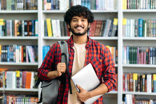 Happy confident indian or arabian male student of university, in stylish casual wear, with backpack and laptop, stands in a library against the background of bookshelves, looks to the camera, smiles photo