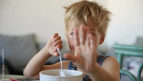 Portrait of cute adorable little blond caucasian sleepy kid boy eating oatmeal porridge early morning. Grumpy child showing stop enough gesture raising hand. Children healthy food nutrition