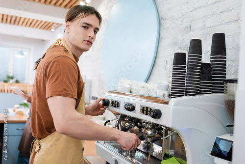Barista in apron looking at camera while making coffee in sweet shop.