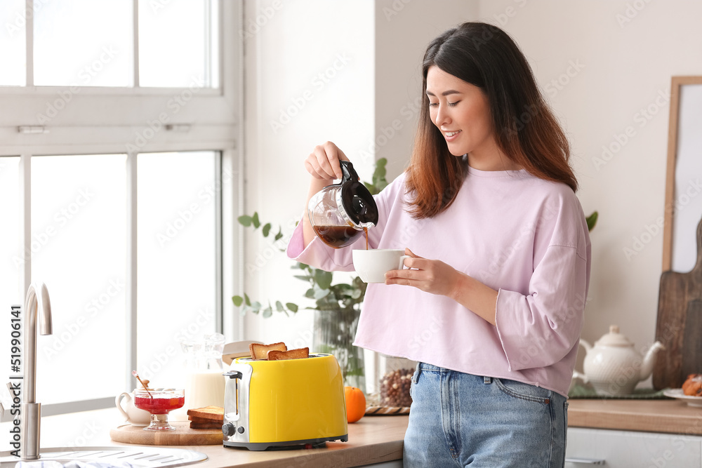Beautiful young Asian woman drinking coffee and making tasty toasts in kitchen Beautiful young Asian woman drinking coffee and making tasty toasts in kitchen