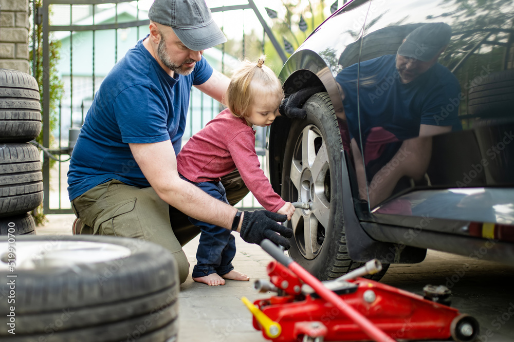 Cute toddler boy helping his father to change car wheels at their backyard. Father teaching his little son to use tools.