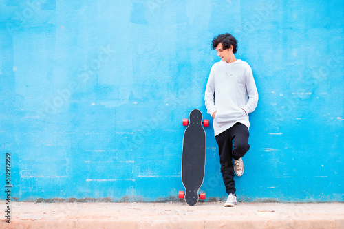 Portrait of a young man with a longboard at his side and blue wall in the background. © Marcio