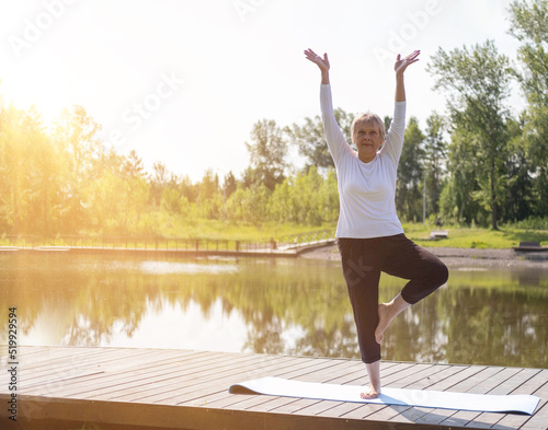 Full-length portrait of a Caucasian elderly woman in yoga vrikshasana poses, tree pose, against the background of a lake in a park. Health and active lifestyle of the elderly.