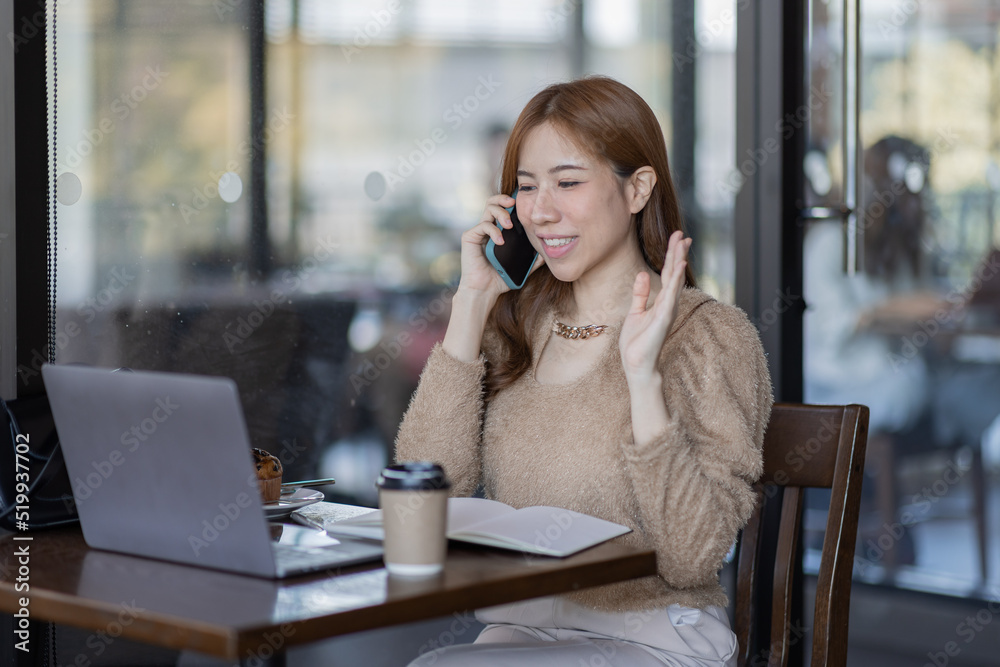 Asian businesswoman conversation on phone using a laptop while sitting in cafe