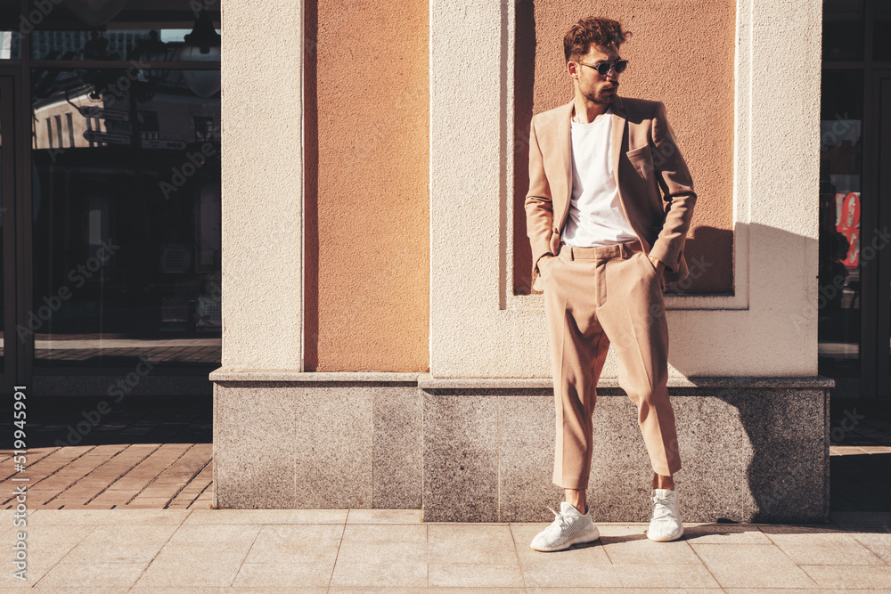 Handsome confident stylish hipster lambersexual model.Sexy modern man dressed in elegant beige suit. Fashion male posing in the street background in Europe city at sunset. In sunglasses. Full length