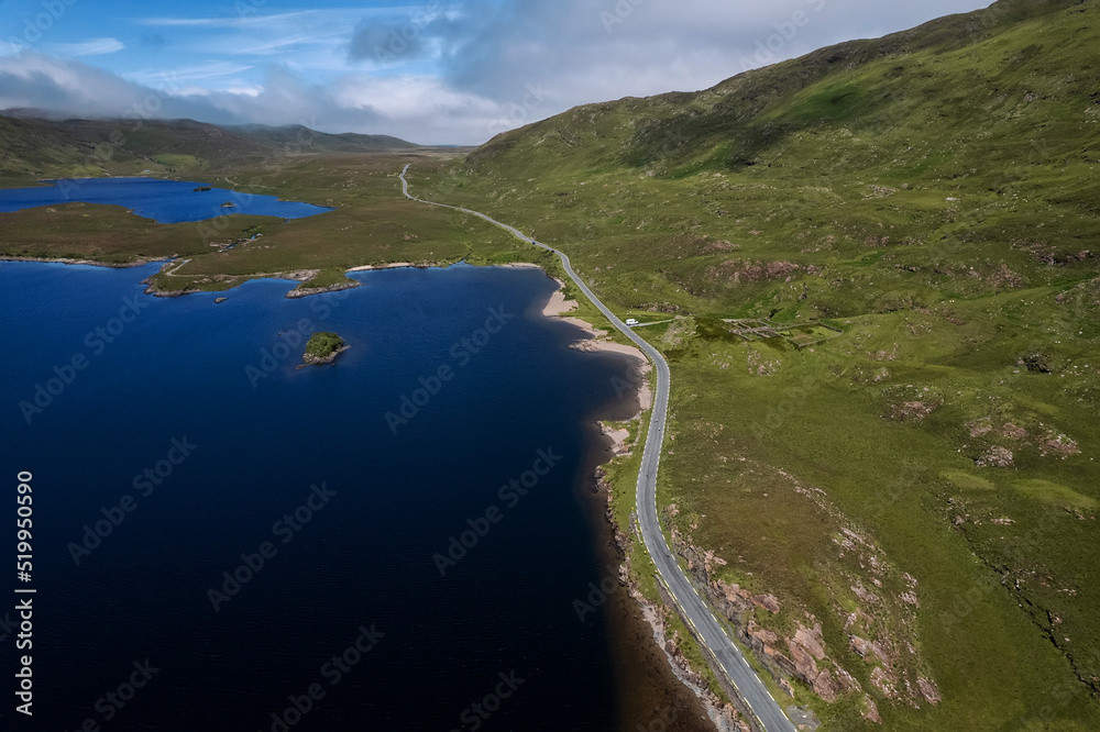 Stunning nature scenery in Connemara, Ireland, Road alongside a big lake with blue water and mountains. Blue cloudy sky over mountain peak. Warm sunny day. Irish landscape. Travel and transport.