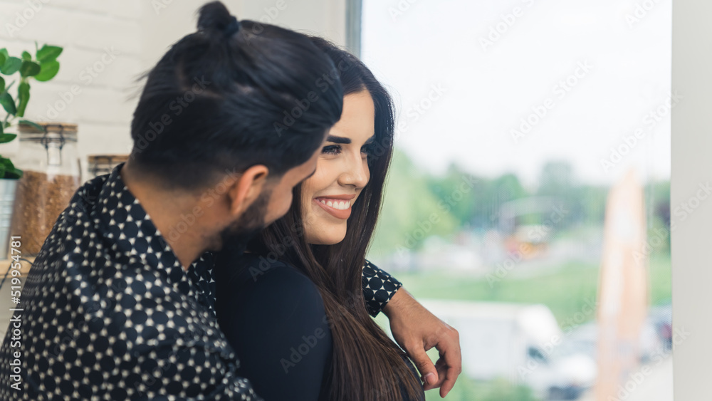 Man with dark hair in a bun embracing his wife smiling standing in the kitchen by the window. Young couple. Indoor shot. High quality photo