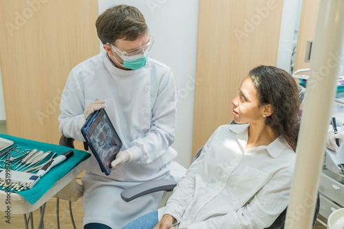 Caucasian male dentist hold digital tablet and explaining tooth problem. Caucasian male doctor using tablet and giving advice or consultation to Latino woman patient about oral care in dental clinic.