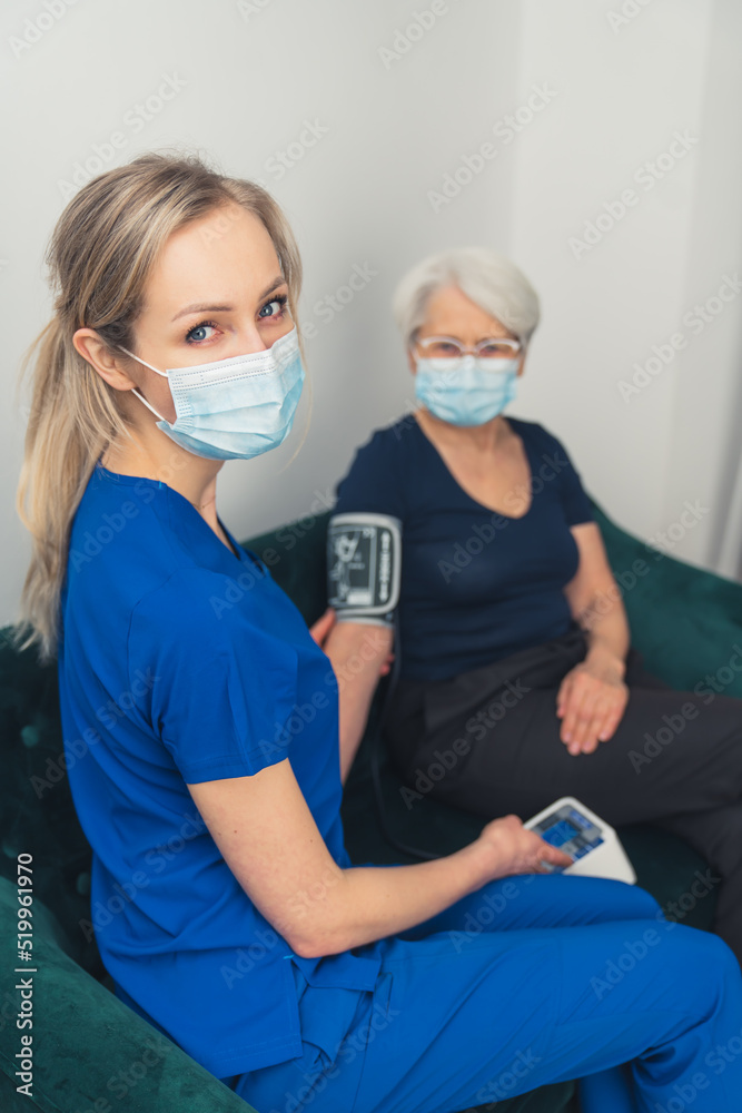 A young, attractive nurse with blue eyes and long blond hair, in a dark blue uniform, measuring the arterial blood pressure of an elderly woman with a face mask on during a pandemic. High-quality