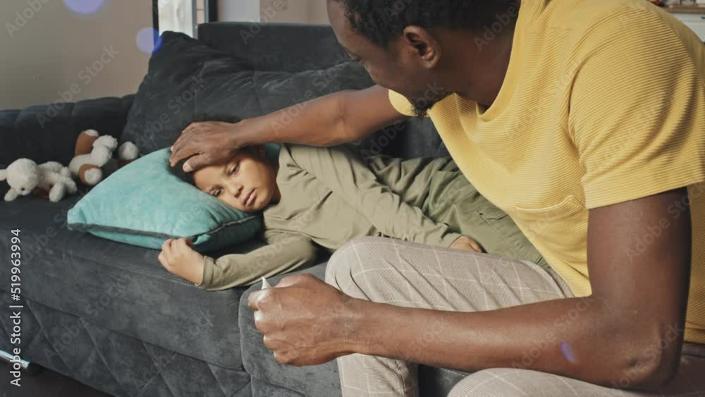 Medium long side view of young African American man sitting on couch, measuring body temperature of sick son Medium long side view of young African American man sitting on couch, measuring body temperature of sick son