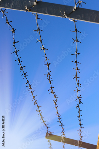 A strip of barbed cold wire in a perishable blue sky. Rays of the sun. Barbed wire fence. Prohibited zone, restriction of freedom.
