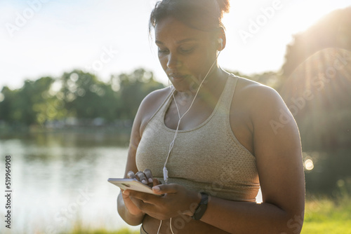 Plus sized African American woman using mobile phone and earphones before exercising at the park in a  summer day photo