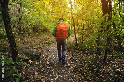 A tourist in a green jacket and a red backpack walks along a mountain path in spring or autumn. Traveler, tourist in autumn landscape. Mountains and trekking, rear view