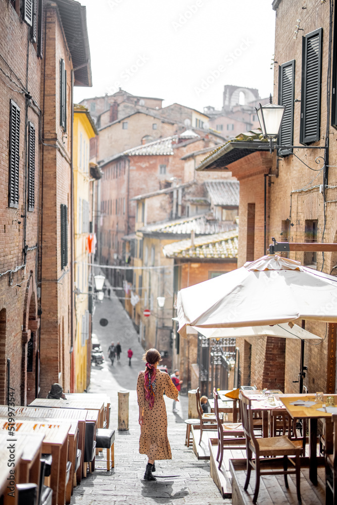 Fototapeta premium Sylish woman walks on narrow and cozy street in old town of Siena city. Concept of traveling in Tuscany region and italian lifestyle
