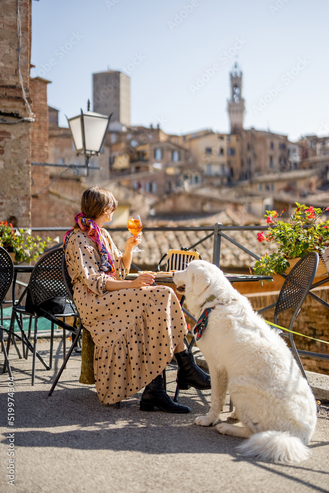 Naklejka premium Woman feeds her dog while sitting at restaurant with beautiful view on Siena town. Traveling Tuscany region in Italy. Concept of italian cuisine