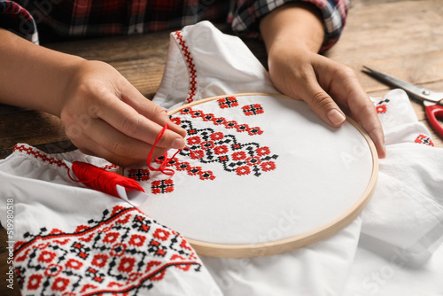Woman embroidering white shirt with colorful threads at wooden table, closeup. Ukrainian national clothes