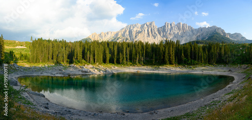 Stunning panoramic view of Carezza Lake (Lago di Carezza) with its emerald green waters, beautiful trees and mountains in the distance during a dramatic sunset.