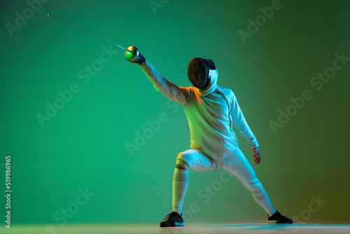 Studio shot of young man, fencer with smallsword practicing fencing isolated on green background in neon light. Sport, energy, skills, achievements