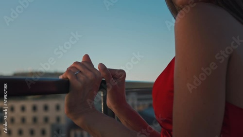 A beautiful woman in a red evening elegant dress stands roof near handrail edge, looks evening city sunset. Party roof luxury hotel. Patio area apartment. Journey new places world. Romantic dinner