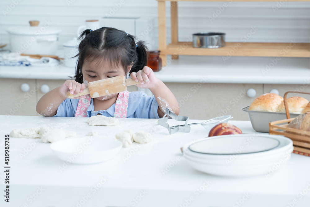 Asian little cute kid girl wear apron with flour mess up on face and arms holding wooden rolling pin kneading dough bread at kitchen home or baking class at school. Child education concept