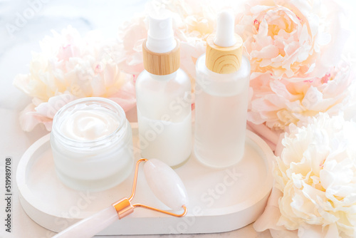 White cosmetic bottle containers with peony flowers on wooden tray. Cosmetics SPA branding mock-up Natural organic beauty product with quartz roller for massage