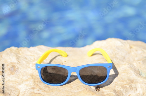 Resort vacation concept - close-up blue yellow sunglasses on stones a bright sunny day by the pool - copy space