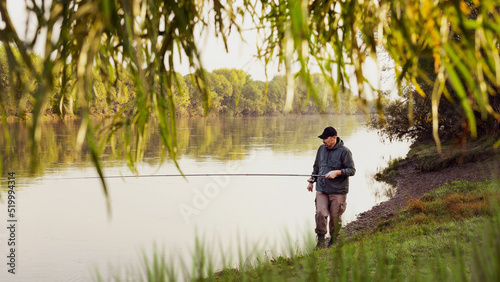 A fisherman in a jacket with a fishing rod stands on the bank of the river. Fishing in the river. Fisherman with a fishing rod on the river bank. Fishing, spinning reel, fish.