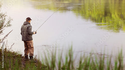 A fisherman in a jacket with a fishing rod stands on the bank of the river. Fishing in the river. Fisherman with a fishing rod on the river bank. Fishing, spinning reel, fish.