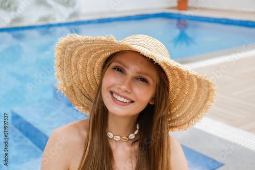 Portrait of smiling fit young woman wearing broad brim straw hat sitting by the pool. Female with Hawaiian puka shell necklace on the neck. Background, copy space, close up. photo