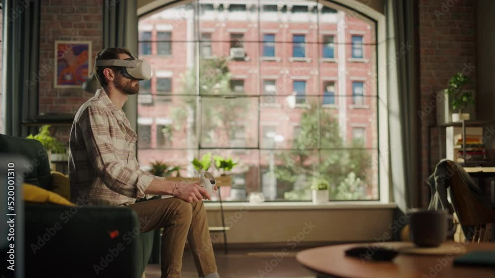 Adult Bearded Male Using Virtual Reality Headset and Controllers at Home. Creative Man Sitting on a Sofa in Living Room, Playing VR Video Game or Working with Special Digital Tools. Adult Bearded Male Using Virtual Reality Headset and Controllers at Home. Creative Man Sitting on a Sofa in Living Room, Playing VR Video Game or Working with Special Digital Tools.