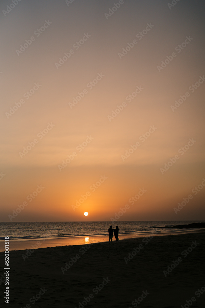 Atardecer en la playa con el sol y horizonte anaranjados con escenas de transeúntes y pescadores