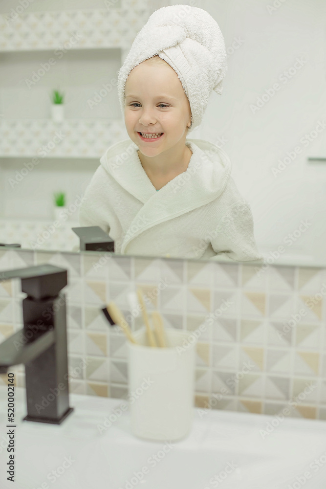 a little girl with a white towel on her head in a white bathrobe in the bathroom looks at her reflection in the mirror a little girl with a white towel on her head in a white bathrobe in the bathroom looks at her reflection in the mirror