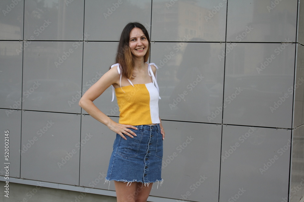 Image of a beautiful stylish woman posing against a dark wall with squares on a summer day in the city. Girl in a knitted crop top and a blue denim skirt in an urban environment. Woman with short bob Image of a beautiful stylish woman posing against a dark wall with squares on a summer day in the city. Girl in a knitted crop top and a blue denim skirt in an urban environment. Woman with short bob