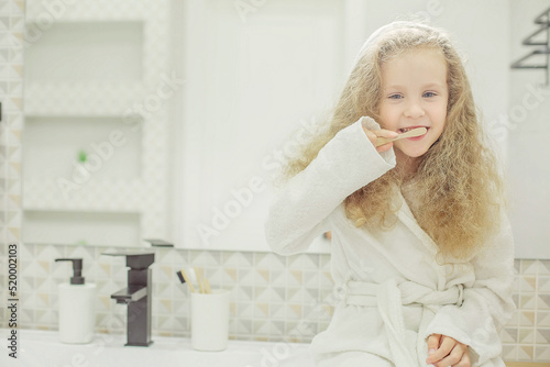 a little girl with long hair in a white bathrobe is holding a wooden toothbrush in the bathroom