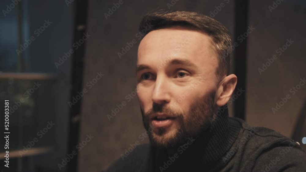 Close-up of bearded Caucasian man in dark gray turtleneck talking to unrecognizable friend indoors at night Close-up of bearded Caucasian man in dark gray turtleneck talking to unrecognizable friend indoors at night