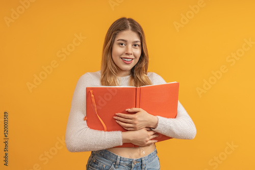 A female student dressed in jeans hugs a textbook, smiles and looks at the camera. On a yellow background. The concept of interesting learning.