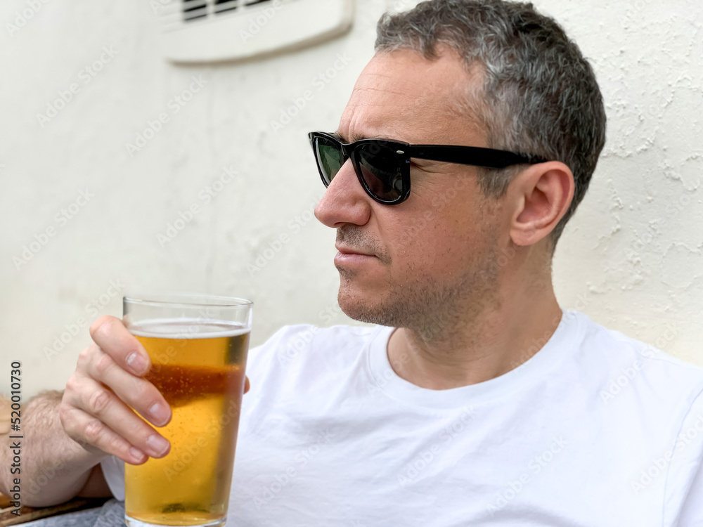 Middle aged man is drinking beer in a cafe or restaurant pub outdoor. Drinking alcohol. Candid. Refreshment, summer time, pub garden. wearing white t-shirt and sunglasses. Drinking lager or ale beer. Middle aged man is drinking beer in a cafe or restaurant pub outdoor. Drinking alcohol. Candid. Refreshment, summer time, pub garden. wearing white t-shirt and sunglasses. Drinking lager or ale beer.