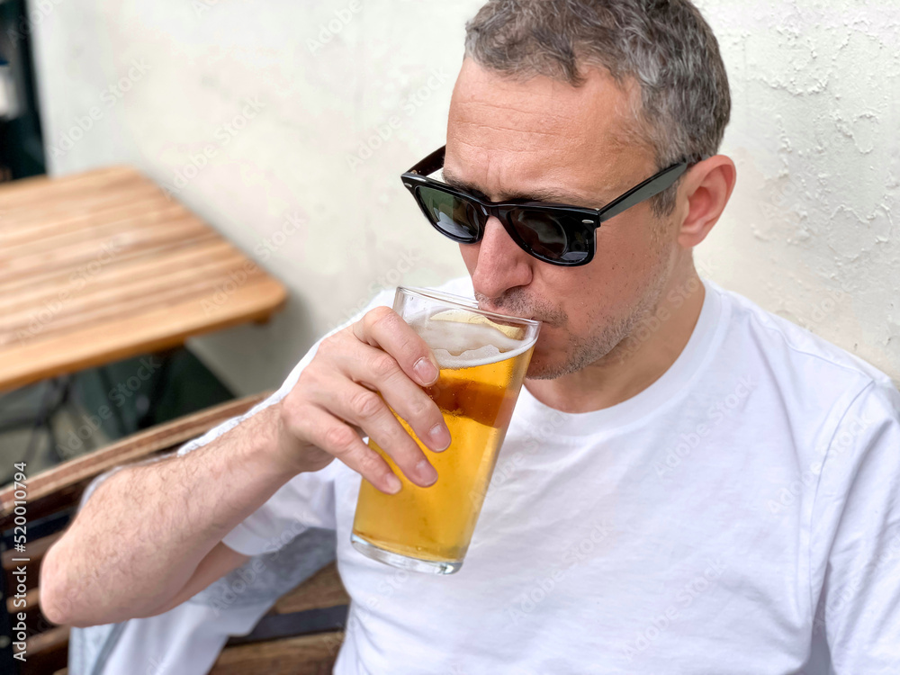 Middle aged man is drinking beer in a cafe or restaurant pub outdoor. Drinking alcohol. Candid. Refreshment, summer time, pub garden. wearing white t-shirt and sunglasses. Drinking lager or ale beer. Middle aged man is drinking beer in a cafe or restaurant pub outdoor. Drinking alcohol. Candid. Refreshment, summer time, pub garden. wearing white t-shirt and sunglasses. Drinking lager or ale beer.