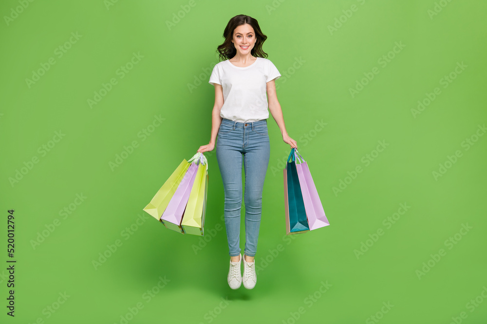 Photo of excited sweet young lady wear white t-shirt jumping high holding shoppers isolated green color background Photo of excited sweet young lady wear white t-shirt jumping high holding shoppers isolated green color background