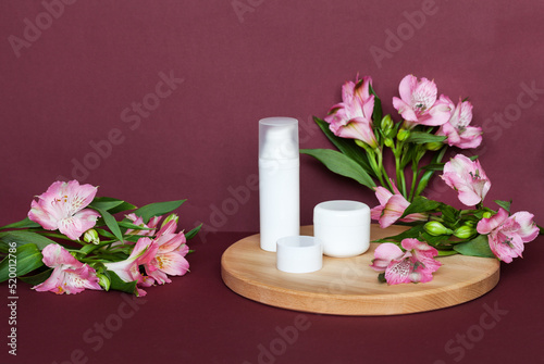 Presentation of summer cosmetic products: anti-aging creams for the face, eyelids and moisturizing gel with vitamin extract on a wooden podium and a burgundy background with alstroemeria flowers