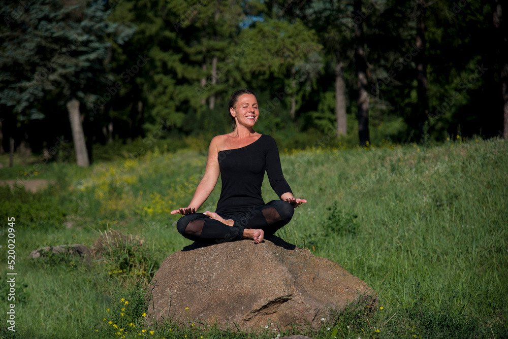 Fototapeta premium young beautiful woman doing yoga exercises on the stone among green grass in the park in sunny day 