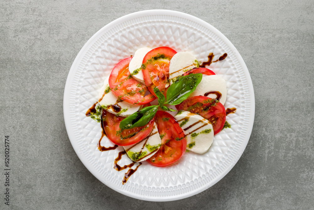Caprese salad with mozzarella and tomato with basil and balsamic and pesto sauce on white plate top view on grey table