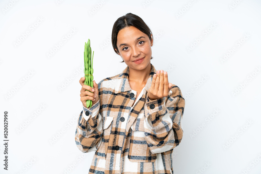 Young hispanic woman holding a green beans isolated on white background inviting to come with hand. Happy that you came Young hispanic woman holding a green beans isolated on white background inviting to come with hand. Happy that you came
