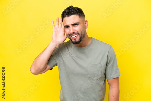 Young Arab handsome man isolated on yellow background listening to something by putting hand on the ear