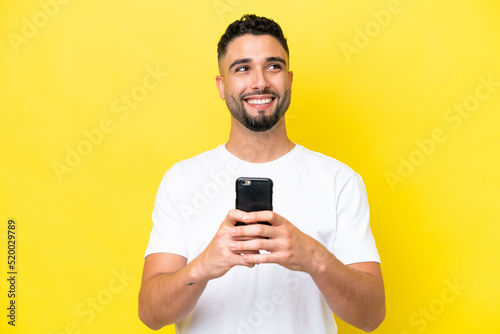 Young Arab handsome man isolated on yellow background using mobile phone and looking up