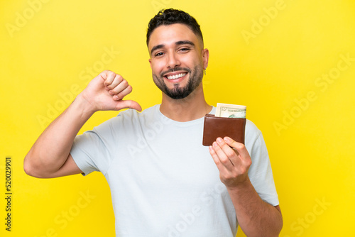 Young Arab man holding a wallet isolated on yellow background proud and self-satisfied