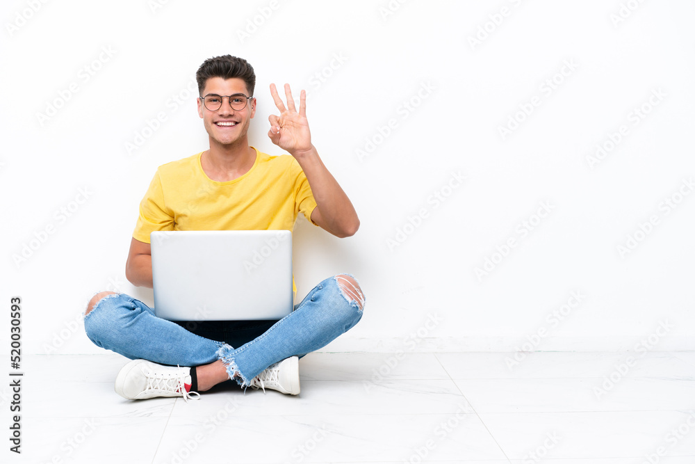 Young man sitting on the floor isolated on white background happy and counting three with fingers Young man sitting on the floor isolated on white background happy and counting three with fingers